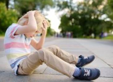 Blonde little boy sitting on sidewalk and holding his head with both of his hands