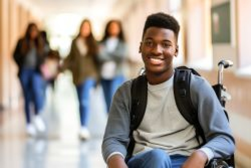 Preteen in a wheelchair smiling with a group of preteen girls in the background walking down a school hallway.